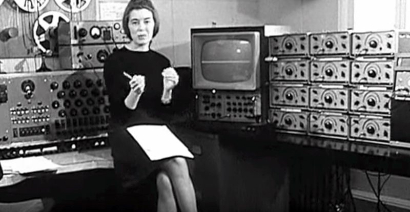 Delia Derbyshire seated among tape machines and oscillators at the BBC Radiophonic Workshop