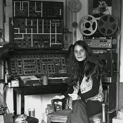 A young Éliane Radigue seated beside her home studio, an ARP 2500 and reel-to-reel tape machines behind her