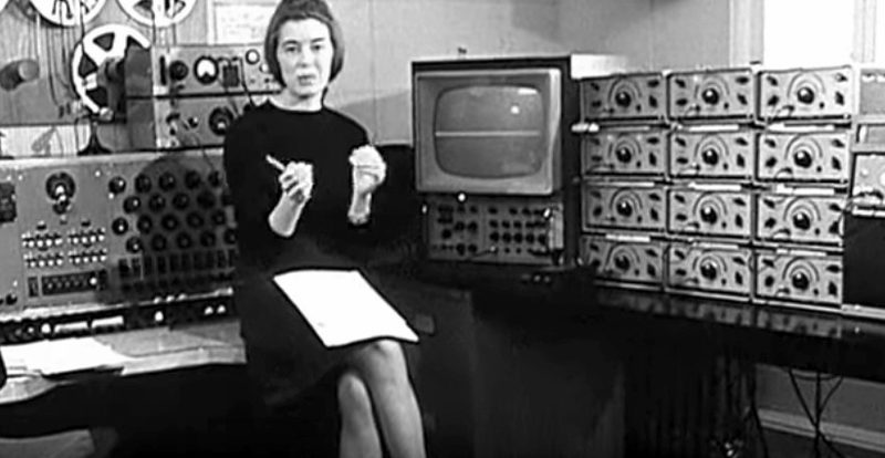 Delia Derbyshire seated among tape machines and oscillators at the BBC Radiophonic Workshop
