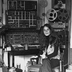 A young Éliane Radigue seated beside her home studio, an ARP 2500 and reel-to-reel tape machines behind her