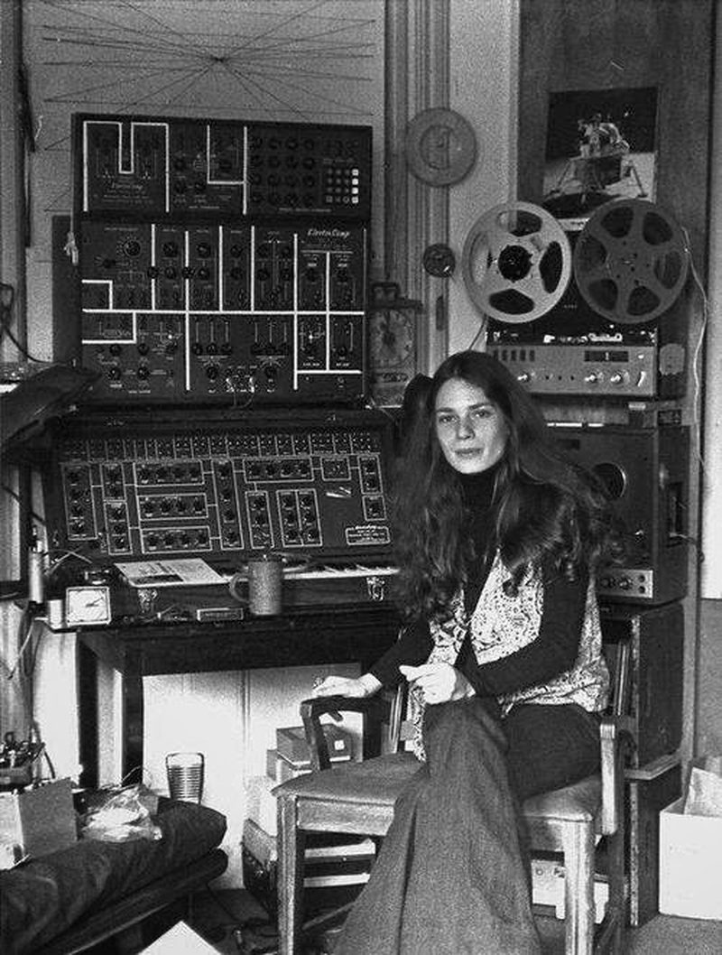 A young Éliane Radigue seated beside her home studio, an ARP 2500 and reel-to-reel tape machines behind her