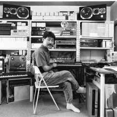 Hiroshi Yoshimura seated at a keyboard in his home studio, shelves of tapes and equipment behind him