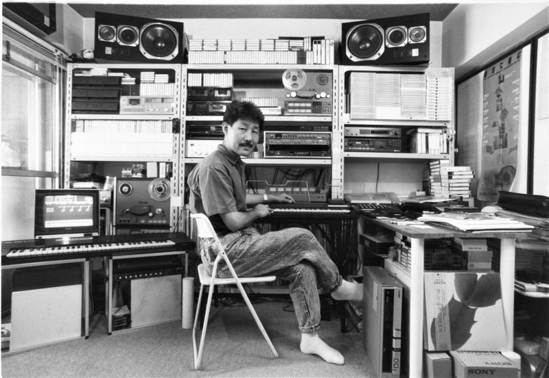 Hiroshi Yoshimura seated at a keyboard in his home studio, shelves of tapes and equipment behind him
