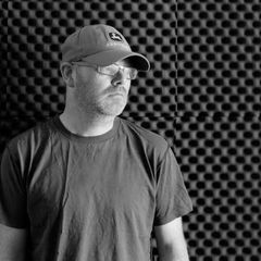 Mark Fell in a cap and glasses against an acoustic-foam wall