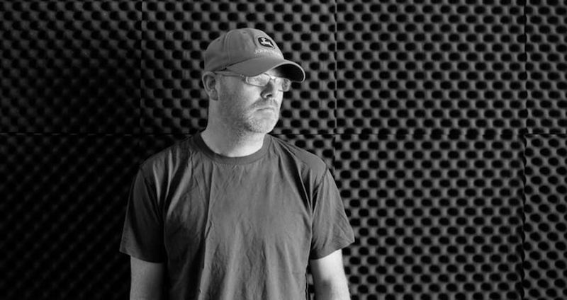 Mark Fell in a cap and glasses against an acoustic-foam wall