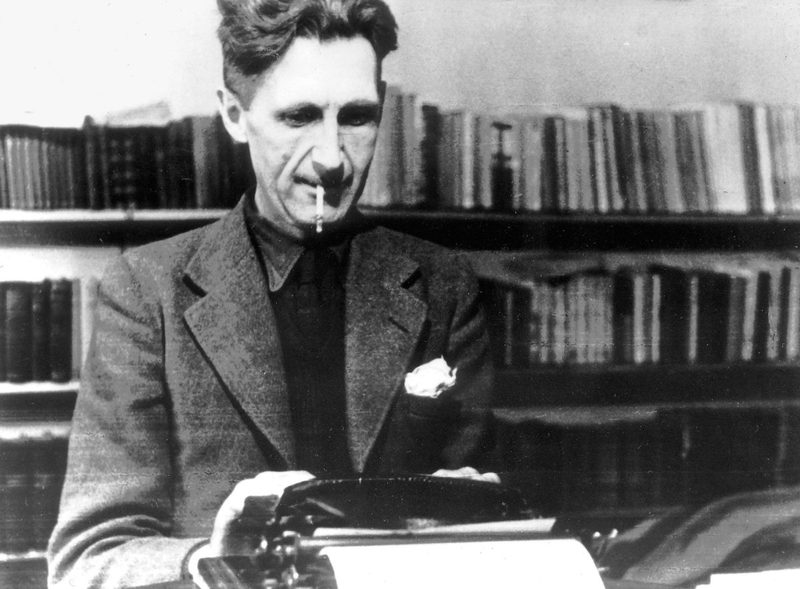 George Orwell typing at a desk, bookshelves behind him