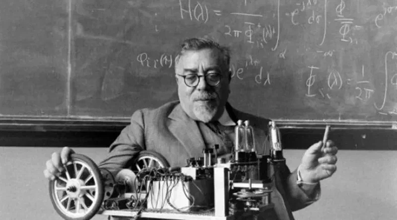 Norbert Wiener holding a cigar in front of a blackboard, with his Palomilla cybernetic moth on the table