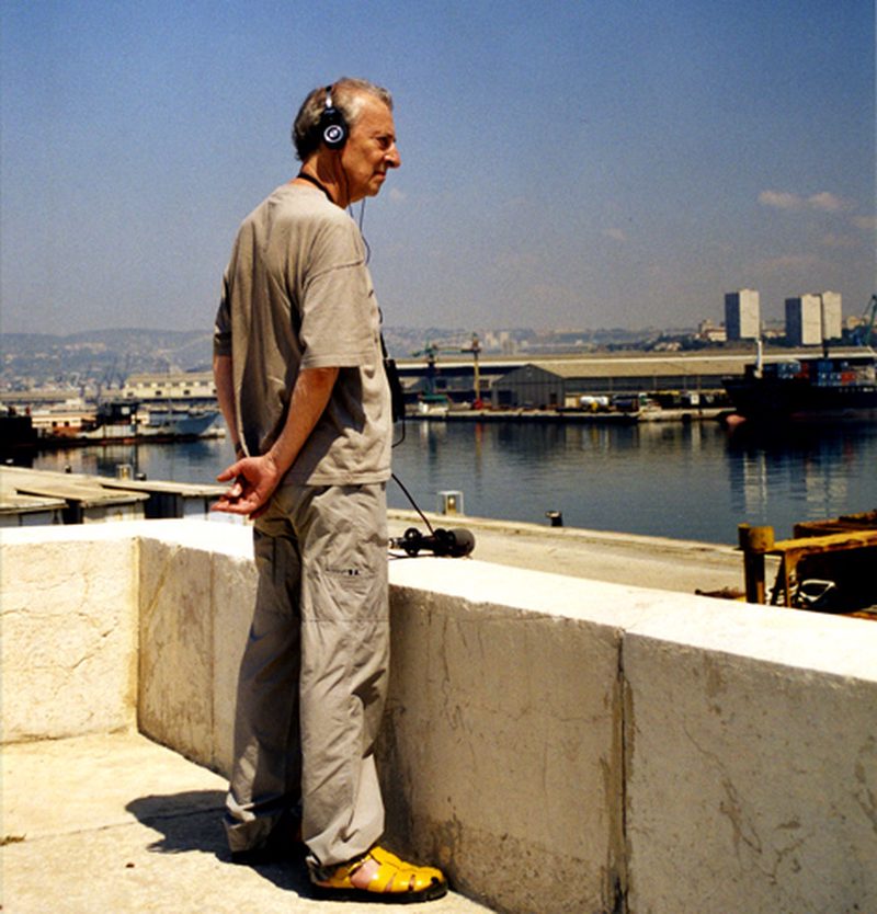 Luc Ferrari wearing headphones with a portable recorder, overlooking a harbor