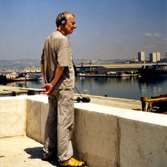 Luc Ferrari wearing headphones with a portable recorder, overlooking a harbor