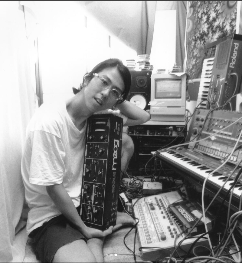Susumu Yokota seated at home holding a Moog Rogue synthesizer, surrounded by studio equipment