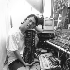 Susumu Yokota seated at home holding a Moog Rogue synthesizer, surrounded by studio equipment