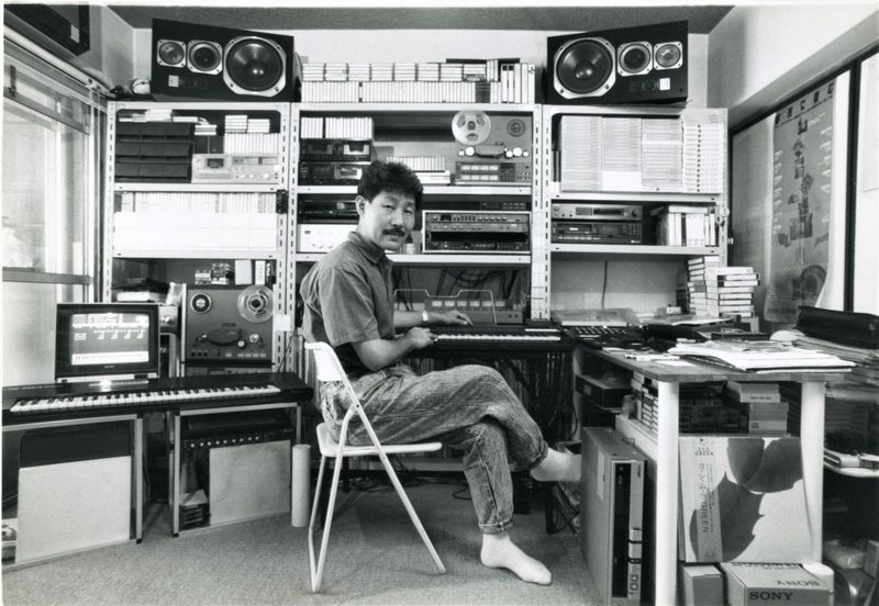 Hiroshi Yoshimura seated at a keyboard in his home studio, shelves of tapes and equipment behind him