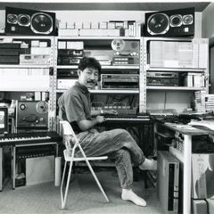 Hiroshi Yoshimura seated at a keyboard in his home studio, shelves of tapes and equipment behind him